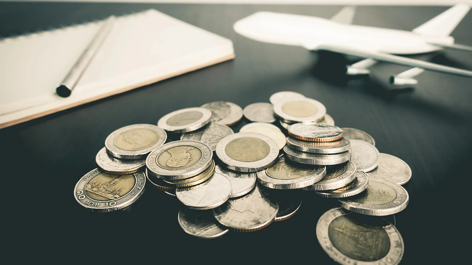 A pile of coins beside a notebook, pen and small toy airplane on a dark table, representing destination wedding budget planning.