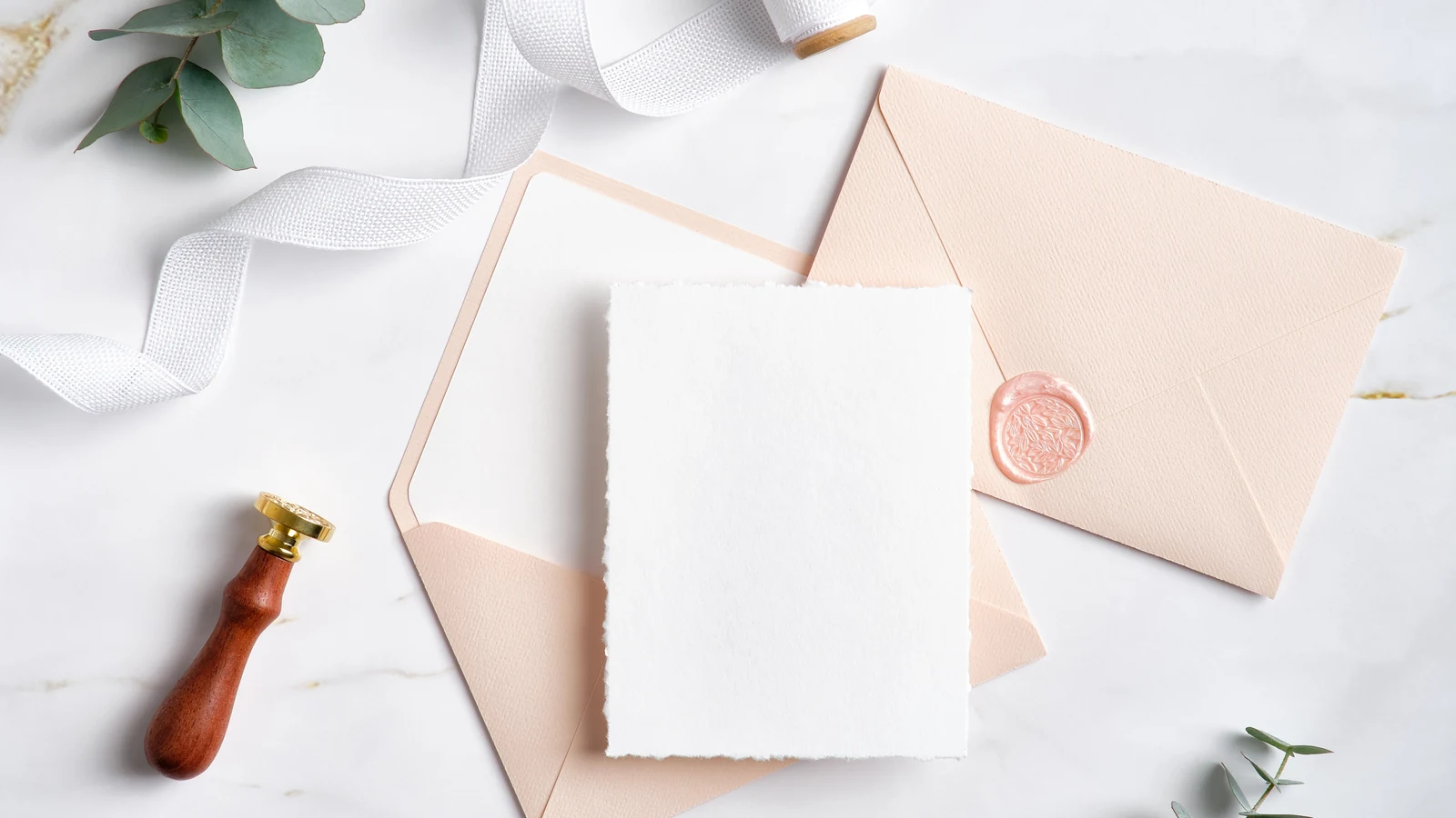A blush envelope, a deckle-edged card and a wax seal on a marble surface.