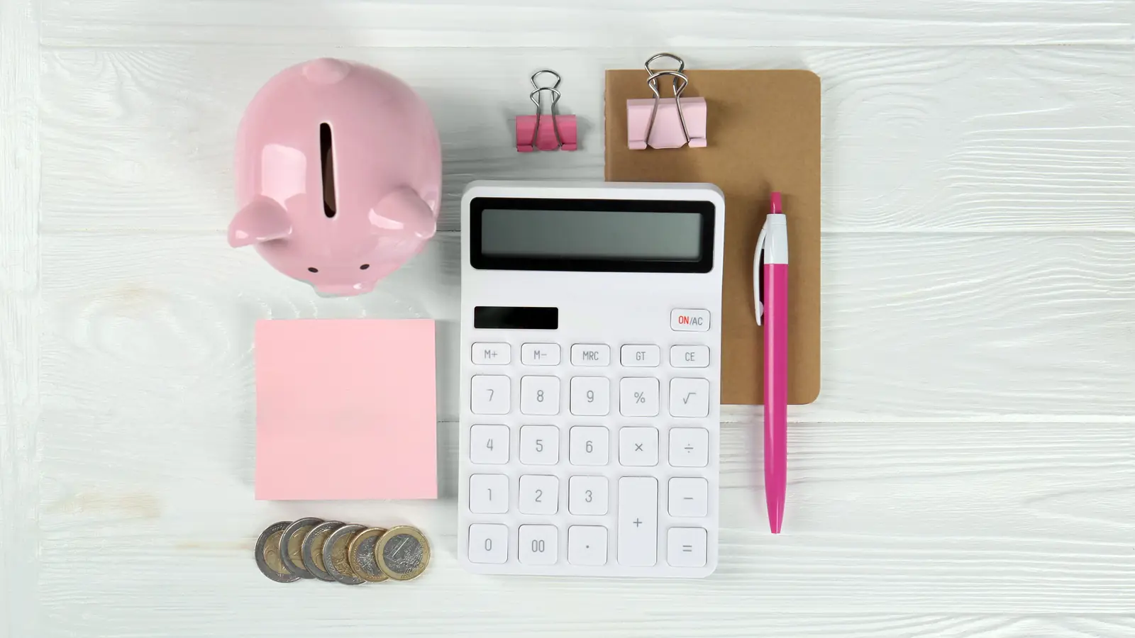 A pink piggy bank, calculator, coins and blank notes arranged on a white tabletop.