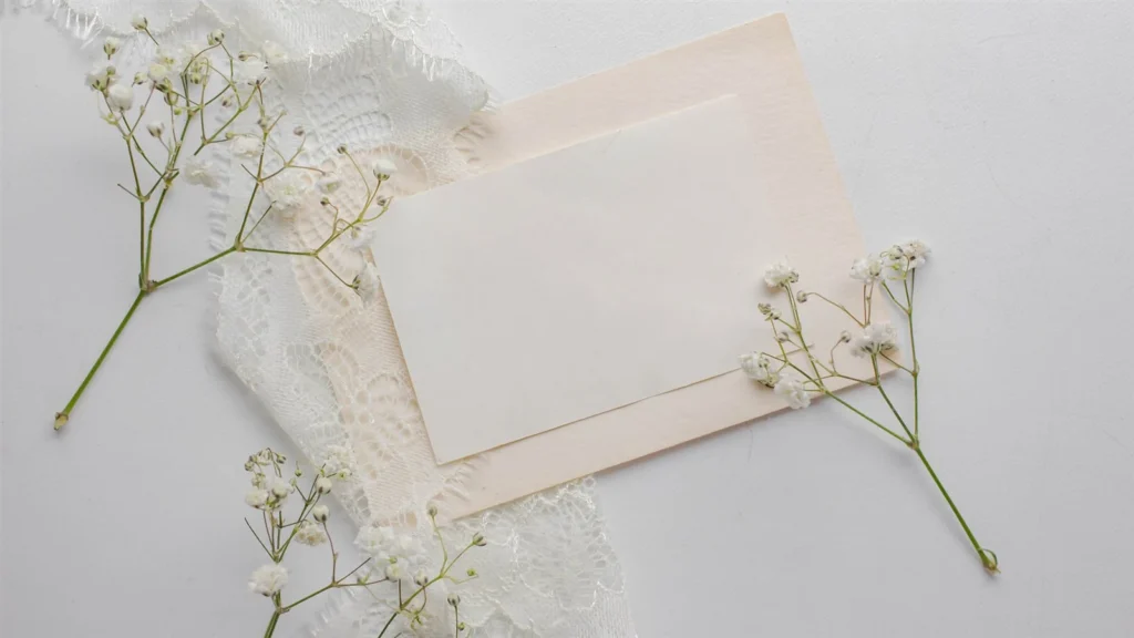 A blank card on a blush envelope, framed by lace and sprigs of gypsophila, on a pale surface.
