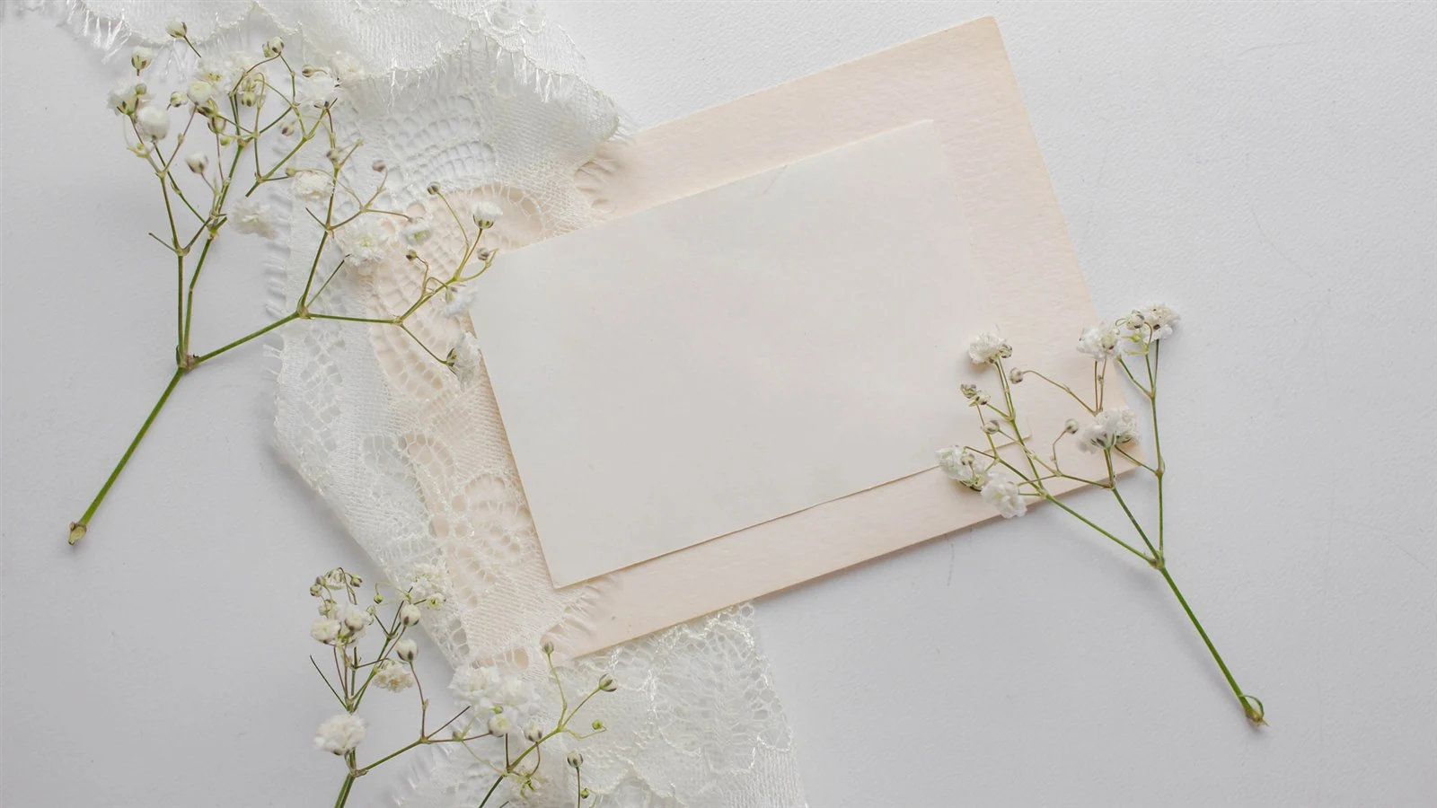 A blank card on a blush envelope, framed by lace and sprigs of gypsophila, on a pale surface.